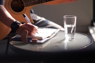 person holding pen writing on white paper beside clear drinking glass