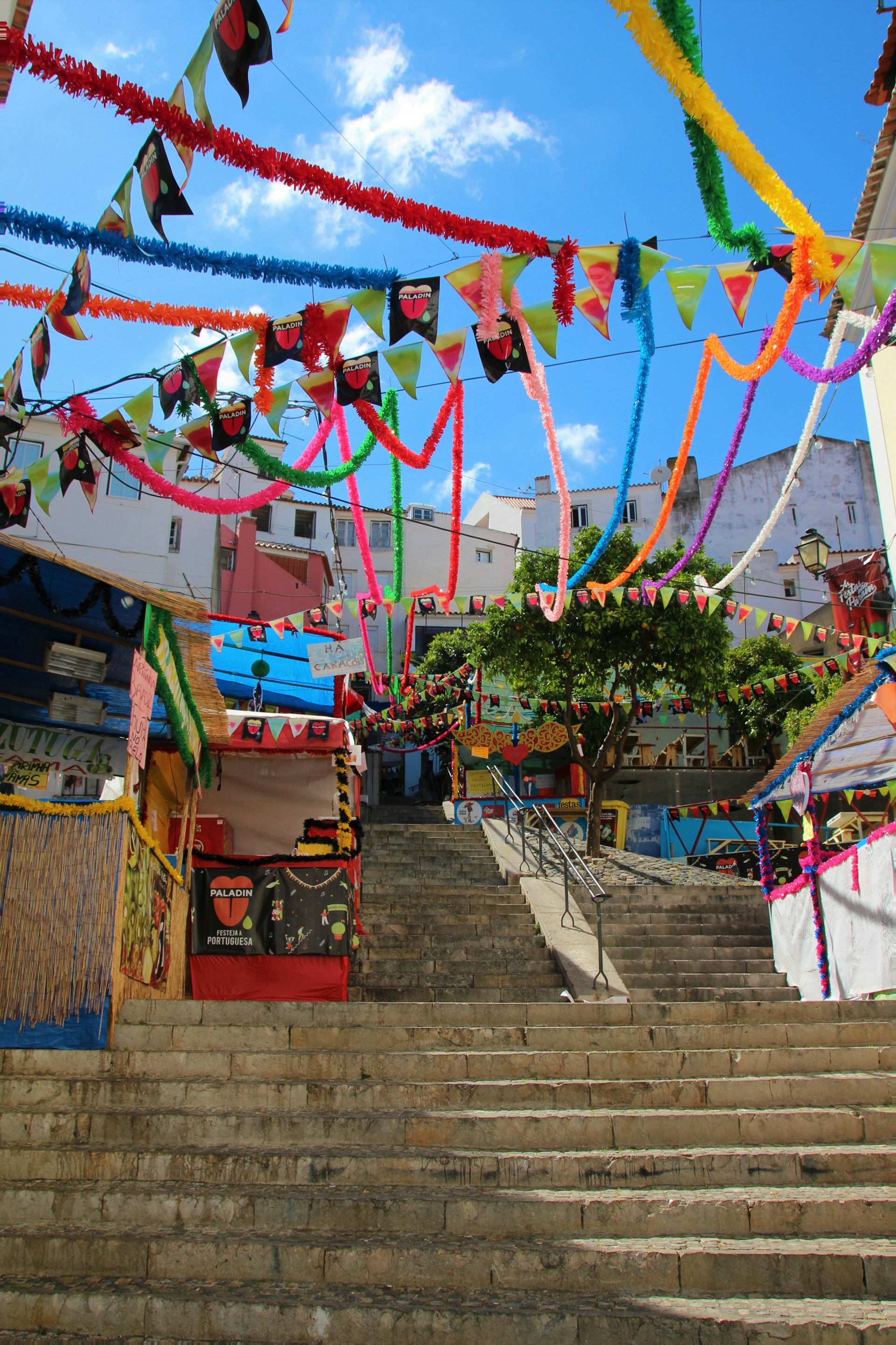 A vibrant and colorful outdoor scene featuring a staircase adorned with a variety of festive decorations. Multicolored streamers and flags hang overhead, creating a lively and cheerful atmosphere. Surrounding buildings are painted in different bright shades, contributing to the festive ambiance, and a tree adds greenery to the mix.