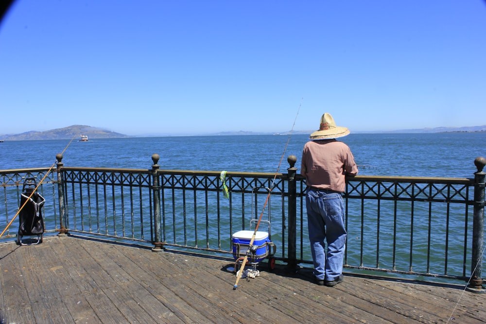 Persona con camisa azul y sombrero marrón de pie sobre barandillas de metal azul cerca del cuerpo de agua