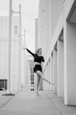 A black and white photograph of a person in a dance pose, gracefully balancing on one foot with the other leg extended. The setting appears to be urban with tall, geometric structures lined up along the sidewalk, creating strong vertical lines.