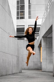 A person is performing a mid-air leap in an alleyway bordered by tall, modern concrete buildings. The subject wears a black long-sleeve top and shorts, with hair flowing dramatically as they jump.