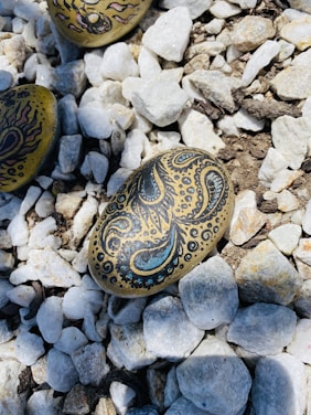 Decorative stones with intricate designs rest on a bed of white and beige pebbles. One stone features a paisley-like pattern in black and blue, surrounded by smaller dots and swirls.