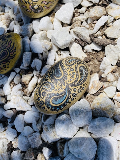 Decorative stones with intricate designs rest on a bed of white and beige pebbles. One stone features a paisley-like pattern in black and blue, surrounded by smaller dots and swirls.