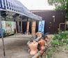 A striped awning shades a group of headless mannequins that are scattered on metal frames near a makeshift storefront. In the background, advertising with images of fresh produce and bottled water can be seen, alongside potted plants and some flowers adding a touch of nature.