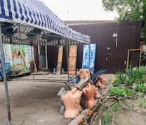 A striped awning shades a group of headless mannequins that are scattered on metal frames near a makeshift storefront. In the background, advertising with images of fresh produce and bottled water can be seen, alongside potted plants and some flowers adding a touch of nature.