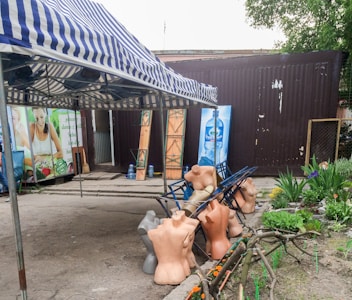 A striped awning shades a group of headless mannequins that are scattered on metal frames near a makeshift storefront. In the background, advertising with images of fresh produce and bottled water can be seen, alongside potted plants and some flowers adding a touch of nature.