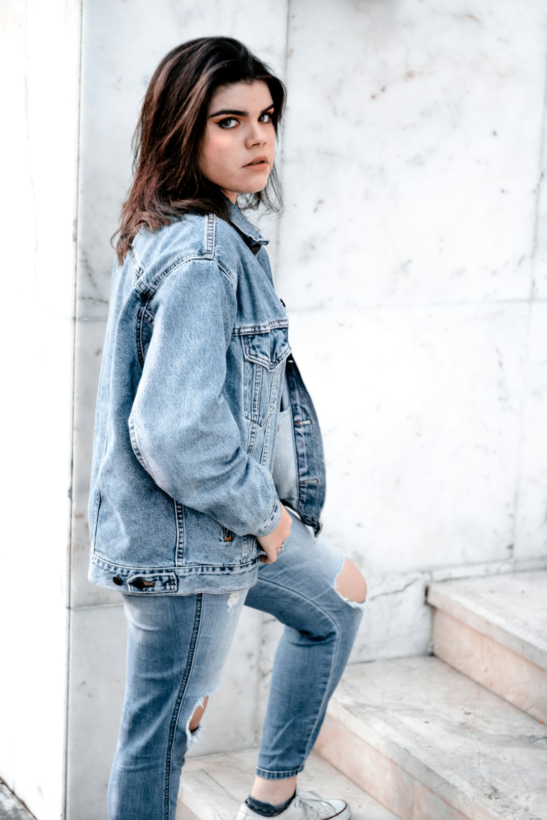 woman in blue denim jacket and blue denim jeans standing beside white wall