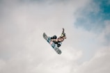 A snowboarder wearing a helmet and goggles performs an aerial trick against a cloudy sky backdrop. Their snowboard has visible branding, and they are captured mid-jump in an action shot.