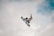 A snowboarder wearing a helmet and goggles performs an aerial trick against a cloudy sky backdrop. Their snowboard has visible branding, and they are captured mid-jump in an action shot.