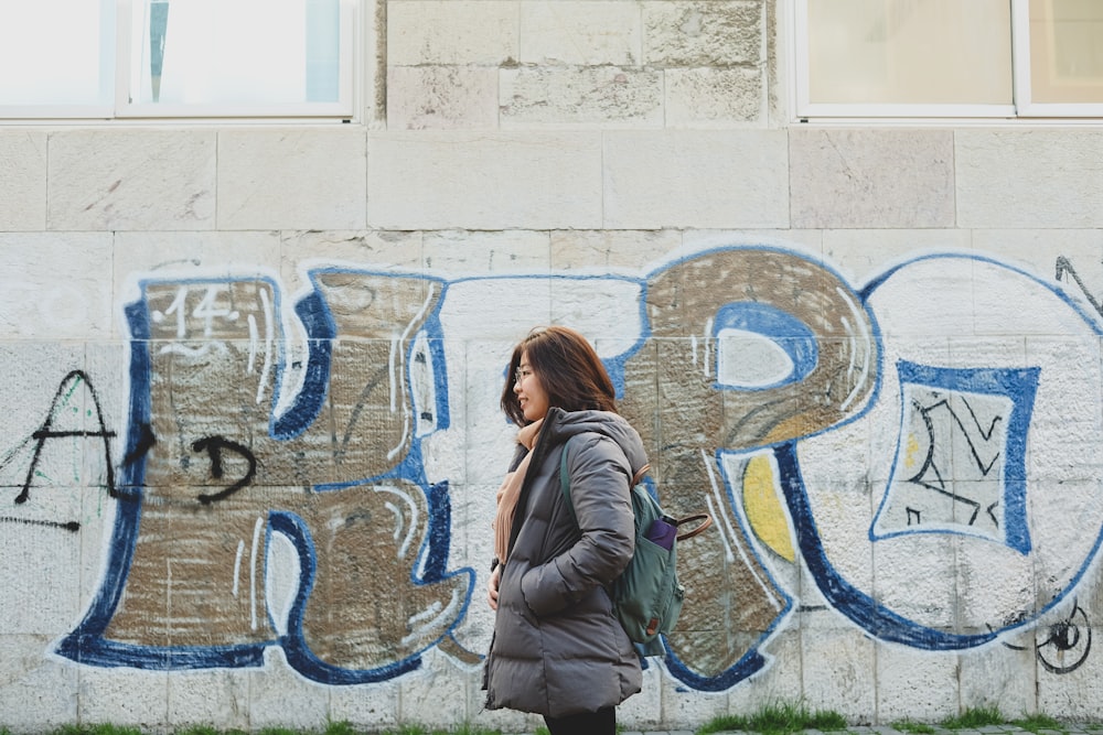 昼間、落書きのある壁のそばに立つ灰色のジャケットを着た女性