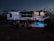 A group of people are gathered outdoors near a large RV camper. The scene is set in the evening with dim lighting. Several individuals sit on folding chairs in a circle, while one person stands at the entrance of the camper. There are lights illuminating parts of the camper, and a small rug is placed on the ground.