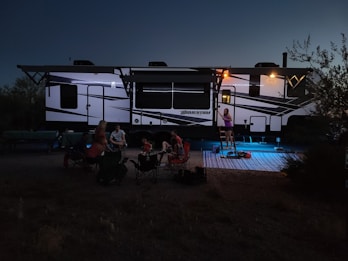A group of people are gathered outdoors near a large RV camper. The scene is set in the evening with dim lighting. Several individuals sit on folding chairs in a circle, while one person stands at the entrance of the camper. There are lights illuminating parts of the camper, and a small rug is placed on the ground.