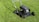 A lawn mower stands on a well-maintained green lawn. The sunlight casts a shadow of the mower, emphasizing its sleek, metallic body and sturdy wheels. Freshly cut grass surrounds the mower, and the bright day adds a vibrant touch to the scene.