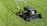A lawn mower stands on a well-maintained green lawn. The sunlight casts a shadow of the mower, emphasizing its sleek, metallic body and sturdy wheels. Freshly cut grass surrounds the mower, and the bright day adds a vibrant touch to the scene.