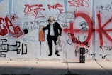 A wall covered in various graffiti, featuring a large painting of a man holding a shopping bag and wearing a suit. Around the figure, there are numerous colorful spray-painted tags, scribbles, and illustrations, including an arrow, a skull with a headdress, and various writings in red, black, and white.