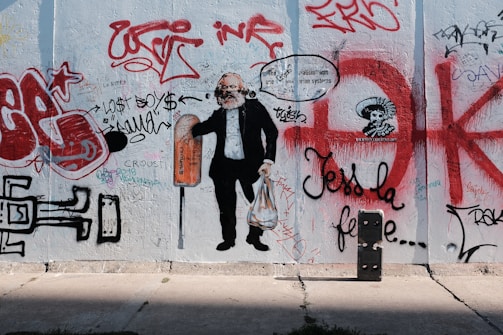 A wall covered in various graffiti, featuring a large painting of a man holding a shopping bag and wearing a suit. Around the figure, there are numerous colorful spray-painted tags, scribbles, and illustrations, including an arrow, a skull with a headdress, and various writings in red, black, and white.