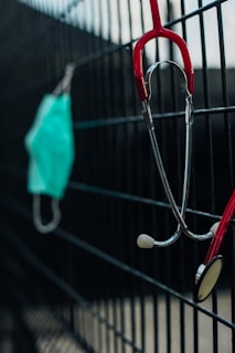 A stethoscope with red tubing is hanging on a black metal fence, and a surgical mask is also hanging next to it slightly blurred in the background.