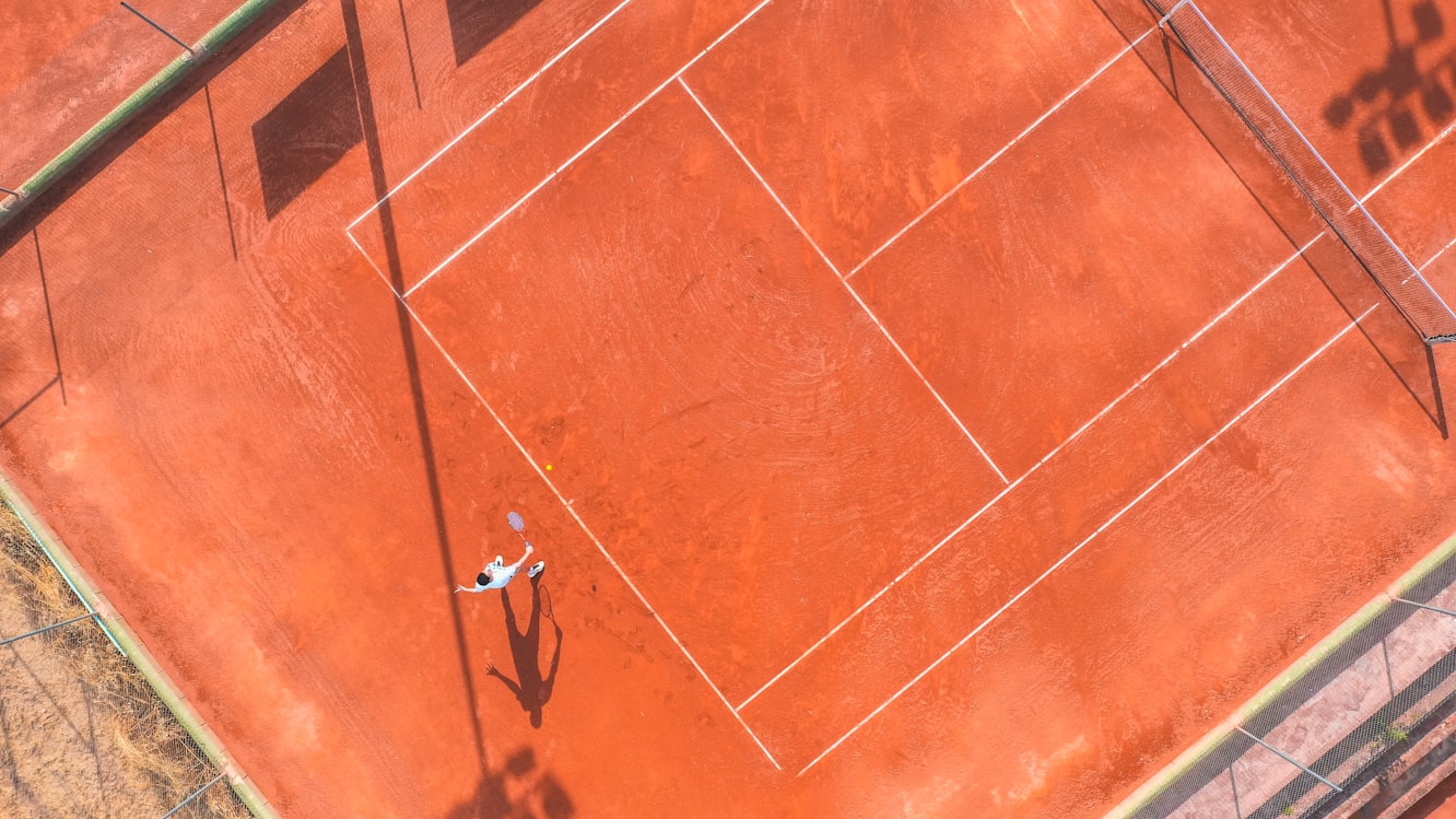 A red clay tennis court with fresh white lines, a net, and a blurred stadium background under bright sunlight