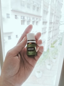 A hand is holding a small bottle labeled 'Stress Away Essential Oil Blend' against a blurred background of a building exterior. The focus is on the bottle, which has a dark green label and white cap.