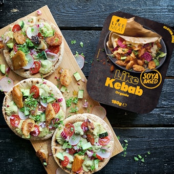 A wooden board holds three pitas topped with a colorful mix of sliced radishes, cucumbers, cherry tomatoes, red onions, grilled soya chunks, and garnished with fresh herbs. Next to the pitas is a package labeled 'Like Kebab', which is described as soya-based and organic. The overall setting suggests a fresh and healthy eating option.