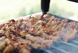 Multiple skewers with marinated pieces of meat being grilled on an outdoor barbecue with smoke rising, giving a sense of freshly cooked food.