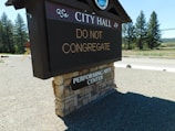 A digital sign in front of a brick-covered base with text reading 'CITY HALL' and 'DO NOT CONGREGATE'. Below, there's a sign indicating a performing arts center. The background features a road, trees, and a clear sky.