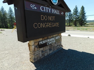 A digital sign in front of a brick-covered base with text reading 'CITY HALL' and 'DO NOT CONGREGATE'. Below, there's a sign indicating a performing arts center. The background features a road, trees, and a clear sky.