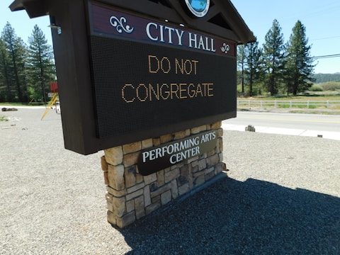 A digital sign in front of a brick-covered base with text reading 'CITY HALL' and 'DO NOT CONGREGATE'. Below, there's a sign indicating a performing arts center. The background features a road, trees, and a clear sky.