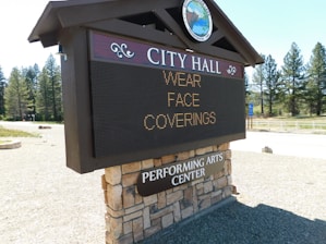 A sign for City Hall displays a message advising to wear face coverings. The structure is made of a combination of stone and wood, and sits outdoors in a natural setting with trees and a clear blue sky in the background. There is also a reference to a Performing Arts Center.