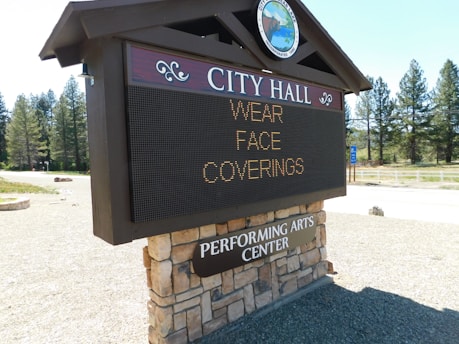 A sign for City Hall displays a message advising to wear face coverings. The structure is made of a combination of stone and wood, and sits outdoors in a natural setting with trees and a clear blue sky in the background. There is also a reference to a Performing Arts Center.