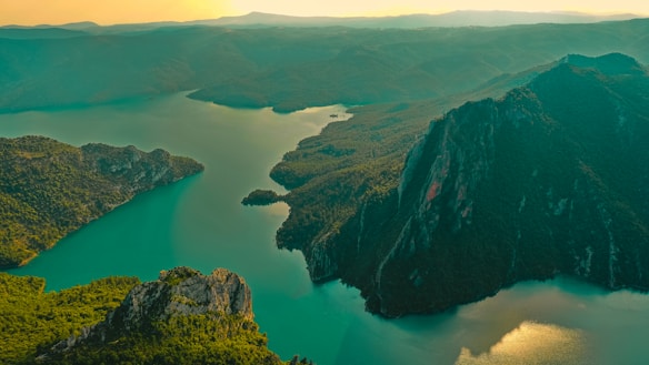 A breathtaking aerial view of a vast landscape featuring a winding river surrounded by lush green hills and dense forests. The river's turquoise waters contrast beautifully with the deep green foliage and rocky cliffs. A hazy, golden light from the sky adds a warm tone to the image, enhancing the natural beauty.