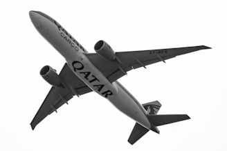 A large commercial cargo airplane in flight, viewed from below against a bright sky. The aircraft has 'Qatar Cargo' written on its body, and it appears to be a twin-engine jet with a distinctive tail design.