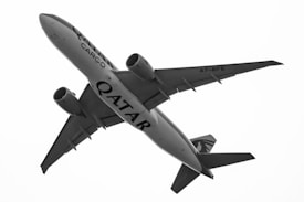 A large commercial cargo airplane in flight, viewed from below against a bright sky. The aircraft has 'Qatar Cargo' written on its body, and it appears to be a twin-engine jet with a distinctive tail design.