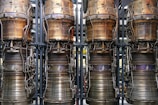 Four large, metallic rocket engines are aligned vertically with intricate piping and components visible. The engines show signs of usage with areas of tarnish and discoloration, emphasizing the industrial and mechanical nature of the objects.