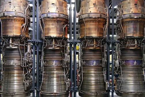 Four large, metallic rocket engines are aligned vertically with intricate piping and components visible. The engines show signs of usage with areas of tarnish and discoloration, emphasizing the industrial and mechanical nature of the objects.