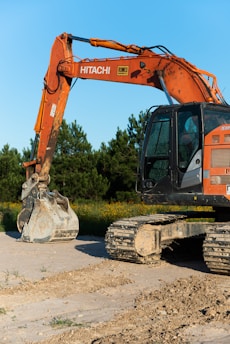 An orange excavator with a large digging bucket stands on a dirt surface. It is positioned in front of a backdrop of green trees under a clear blue sky. The machine shows signs of use, with dirt on its tracks and bucket.