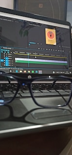 A pair of glasses is resting on a laptop keyboard. The laptop screen displays video editing software with a project timeline and a small image thumbnail labeled 'Sound On Podcast'.