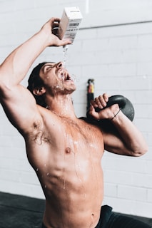 A shirtless man is in a gym setting, pouring water from a carton into his mouth with water splashing down his chest. He is holding a kettlebell on his shoulder with his other hand, and his body is wet with water.