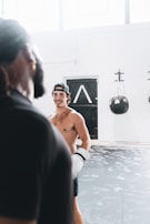 A shirtless person wearing boxing gloves is in an indoor gym setting, appearing engaged with another person in the foreground. The room is well-lit with several windows, and a black punching bag is visible against the white walls. The floor is covered with patterned mats.