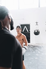 A shirtless person wearing boxing gloves is in an indoor gym setting, appearing engaged with another person in the foreground. The room is well-lit with several windows, and a black punching bag is visible against the white walls. The floor is covered with patterned mats.