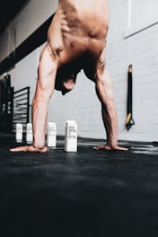 A shirtless person performing a handstand on a gym floor, with several cartons reading 'Boxed Water Is Better' positioned in the background. The environment features gym equipment, and the lighting highlights the muscular build of the person.