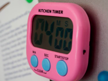 A digital kitchen timer with a pink casing is attached to a metallic surface. The timer displays '04:00' and features blue buttons labeled 'MIN', 'SEC', and 'START/STOP'. The word 'KITCHEN TIMER' is printed in black above the display.
