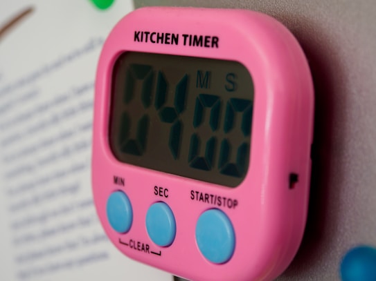 A digital kitchen timer with a pink casing is attached to a metallic surface. The timer displays '04:00' and features blue buttons labeled 'MIN', 'SEC', and 'START/STOP'. The word 'KITCHEN TIMER' is printed in black above the display.