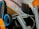 A collection of kitchen utensils is arranged in a holder, including a pair of scissors with blue and orange handles, a metal spoon, and several metal straws or brush handles. The items appear to be both metal and plastic, clustered together with a focus on utility.