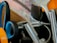 A collection of kitchen utensils is arranged in a holder, including a pair of scissors with blue and orange handles, a metal spoon, and several metal straws or brush handles. The items appear to be both metal and plastic, clustered together with a focus on utility.