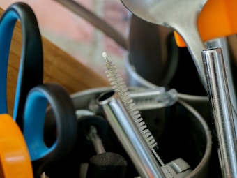 A collection of kitchen utensils is arranged in a holder, including a pair of scissors with blue and orange handles, a metal spoon, and several metal straws or brush handles. The items appear to be both metal and plastic, clustered together with a focus on utility.