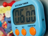 A digital kitchen timer with a blue casing and large black numbers displayed. Below the display, it has two large orange buttons labeled 'MIN' and 'SEC', and a smaller text 'START/STOP'. In the background, a blurred image of a comic book page is visible.