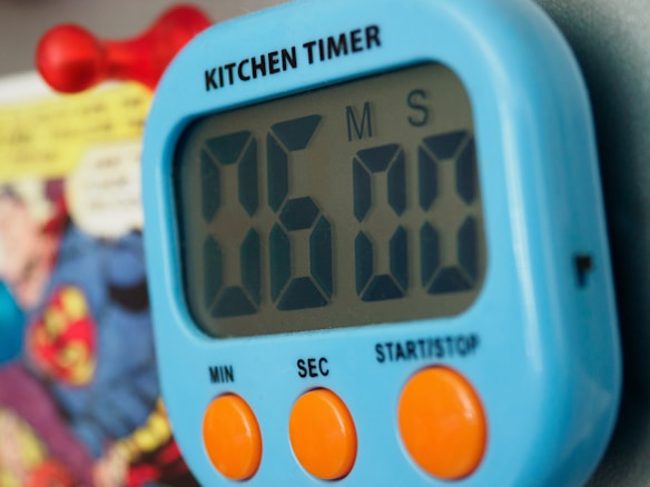 A digital kitchen timer with a blue casing and large black numbers displayed. Below the display, it has two large orange buttons labeled 'MIN' and 'SEC', and a smaller text 'START/STOP'. In the background, a blurred image of a comic book page is visible.