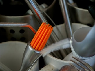 A close-up of various kitchen utensils in a holder. There are metal spoons, forks, and a brush visible. A group of bright orange chopsticks stands out prominently among the utensils.