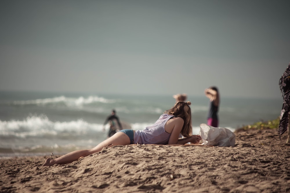 woman in blue bikini lying on brown sand near body of water during daytime